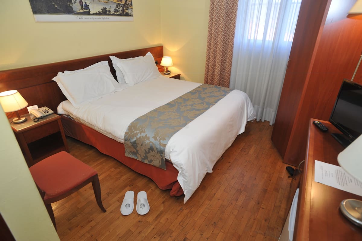A hotel room with a bed, bedside table with a lamp, a chair, slippers, a TV, and a desk with a remote control.