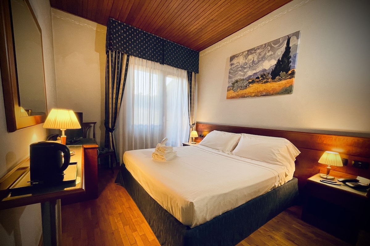 A hotel room with a wooden floor, a bed with white linens, a painting on the wall, a desk with a lamp, and a coffee maker.