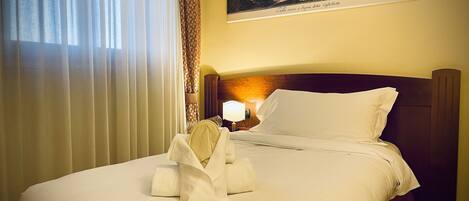 A hotel room with a neatly made bed, a painting on the wall, and a window with curtains.
