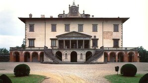 Un grande edificio storico con un ingresso centrale affiancato da archi, una facciata simmetrica e un vialetto sterrato che conduce all'ingresso.