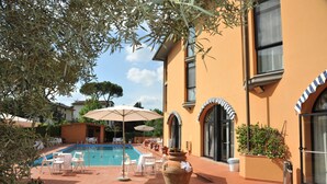 Un'area piscina con lettini e ombrelloni, un grande vaso di fiori e un edificio con finestre ad arco e un tendone a righe.