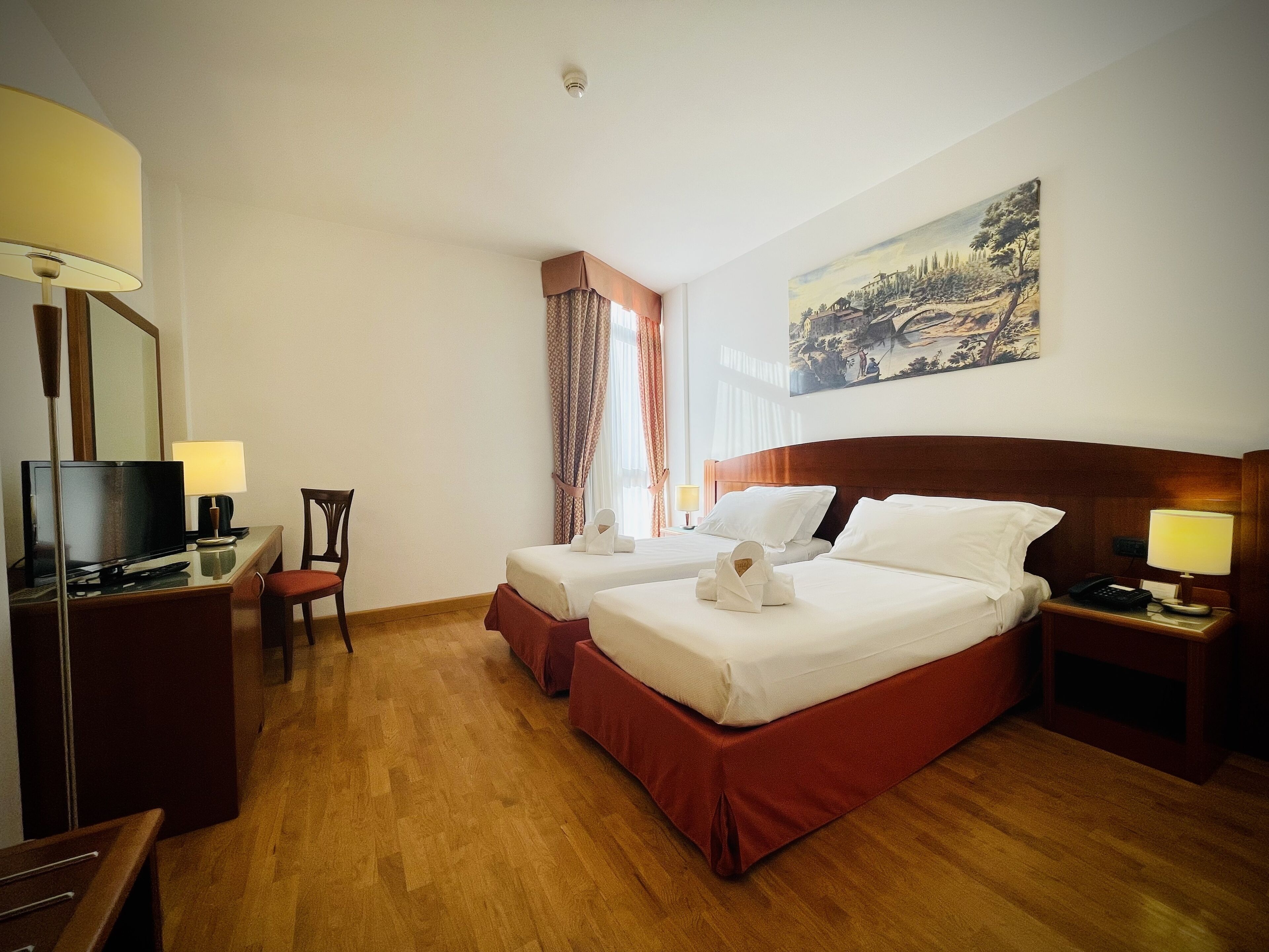Ein Hotelzimmer mit zwei Betten, einem Schreibtisch mit Fernseher, einem Sessel, einer Schreibtischlampe, einem Nachttisch mit Lampe und einem Bild an der Wand.