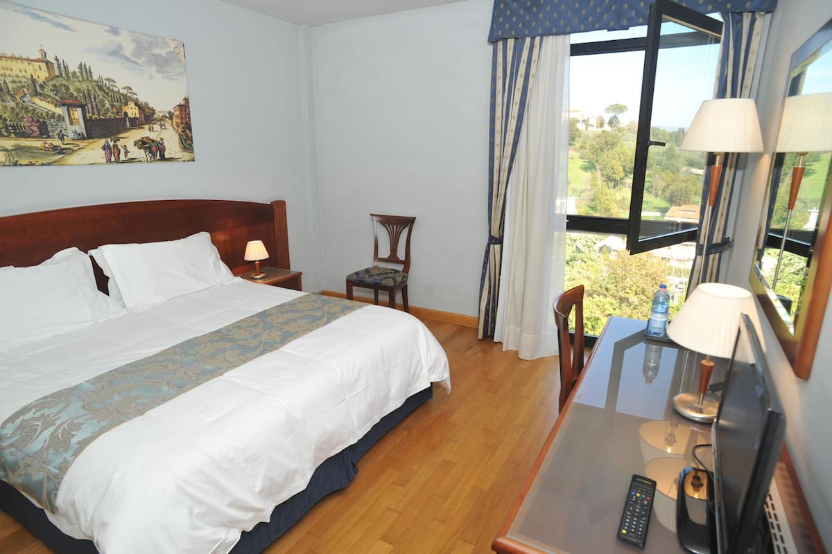 A hotel room with a bed, a chair, a table with a lamp, a remote control, and a painting on the wall.