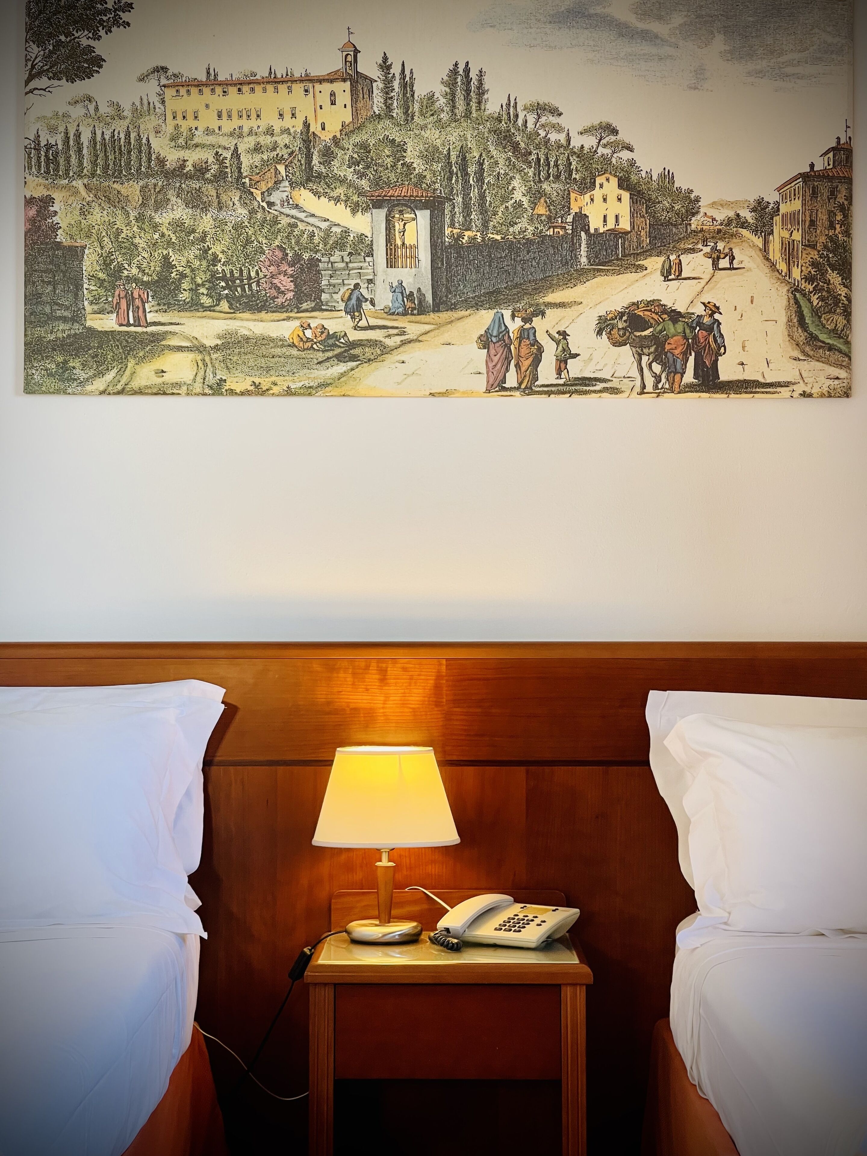 A hotel room with a bed, a bedside table with a lamp and a phone, and a painting of a village scene on the wall.