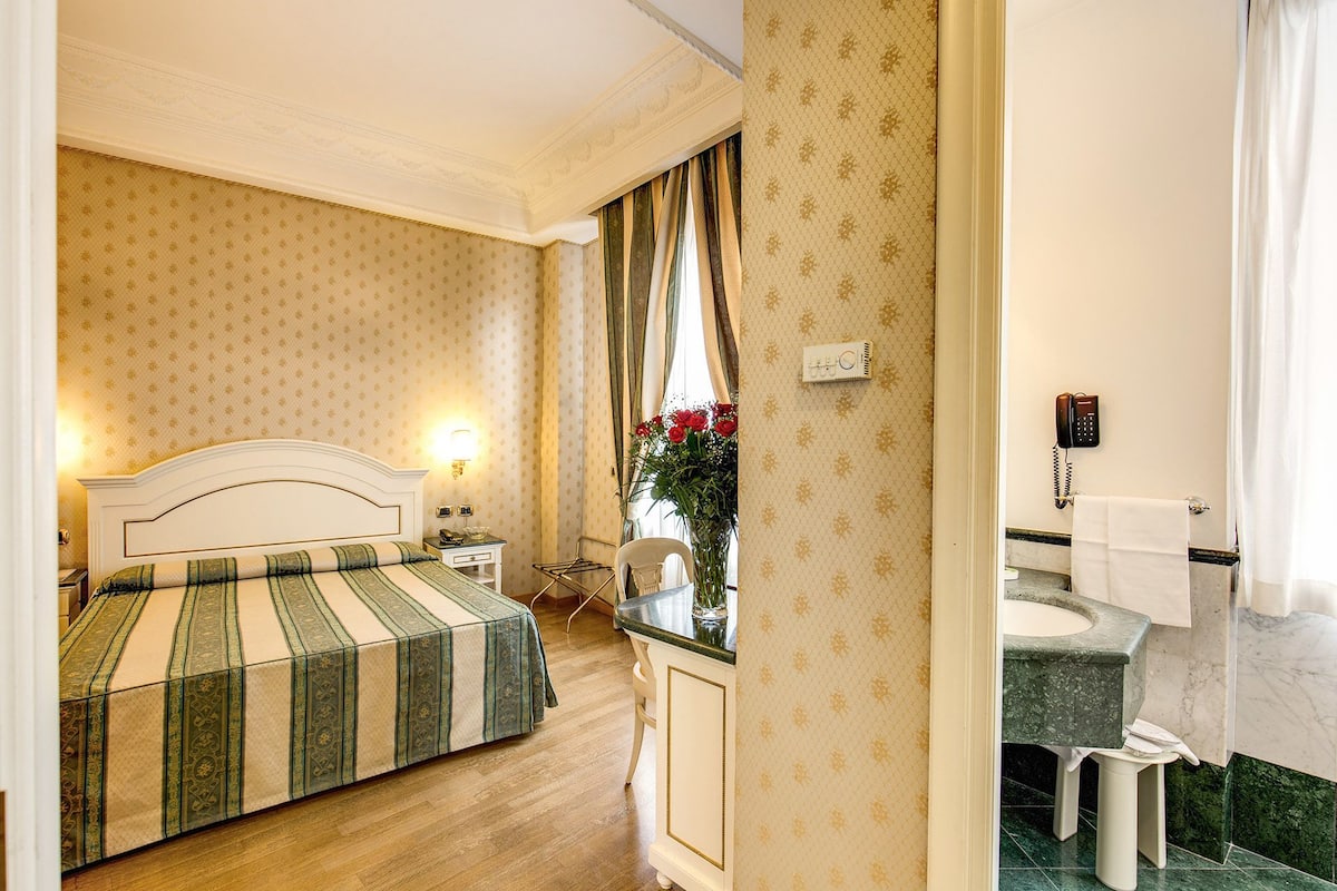 Une chambre d’hôtel comprenant un lit, une table de chevet, un bureau, une chaise, un vase avec des fleurs, un téléphone et une salle de bain avec un lavabo en marbre.