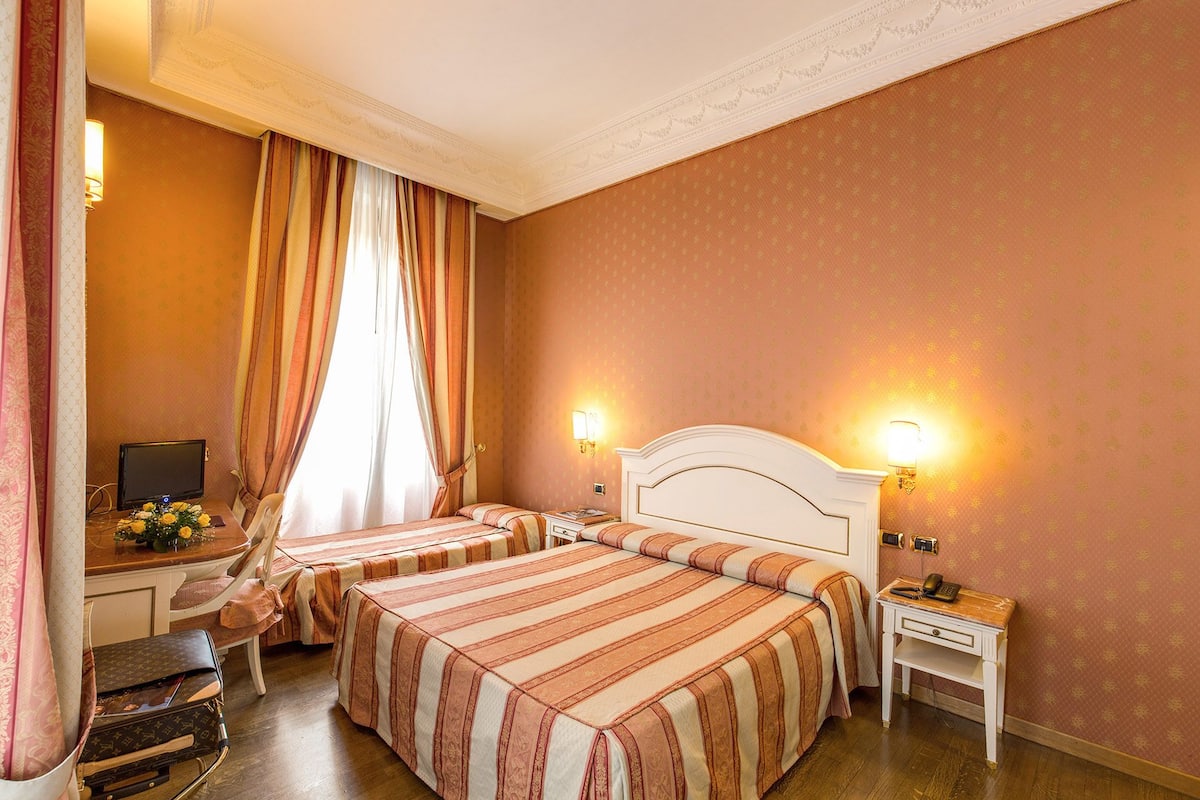 Une chambre d’hôtel avec deux lits, un bureau avec un ordinateur et une table de chevet avec une lampe.
