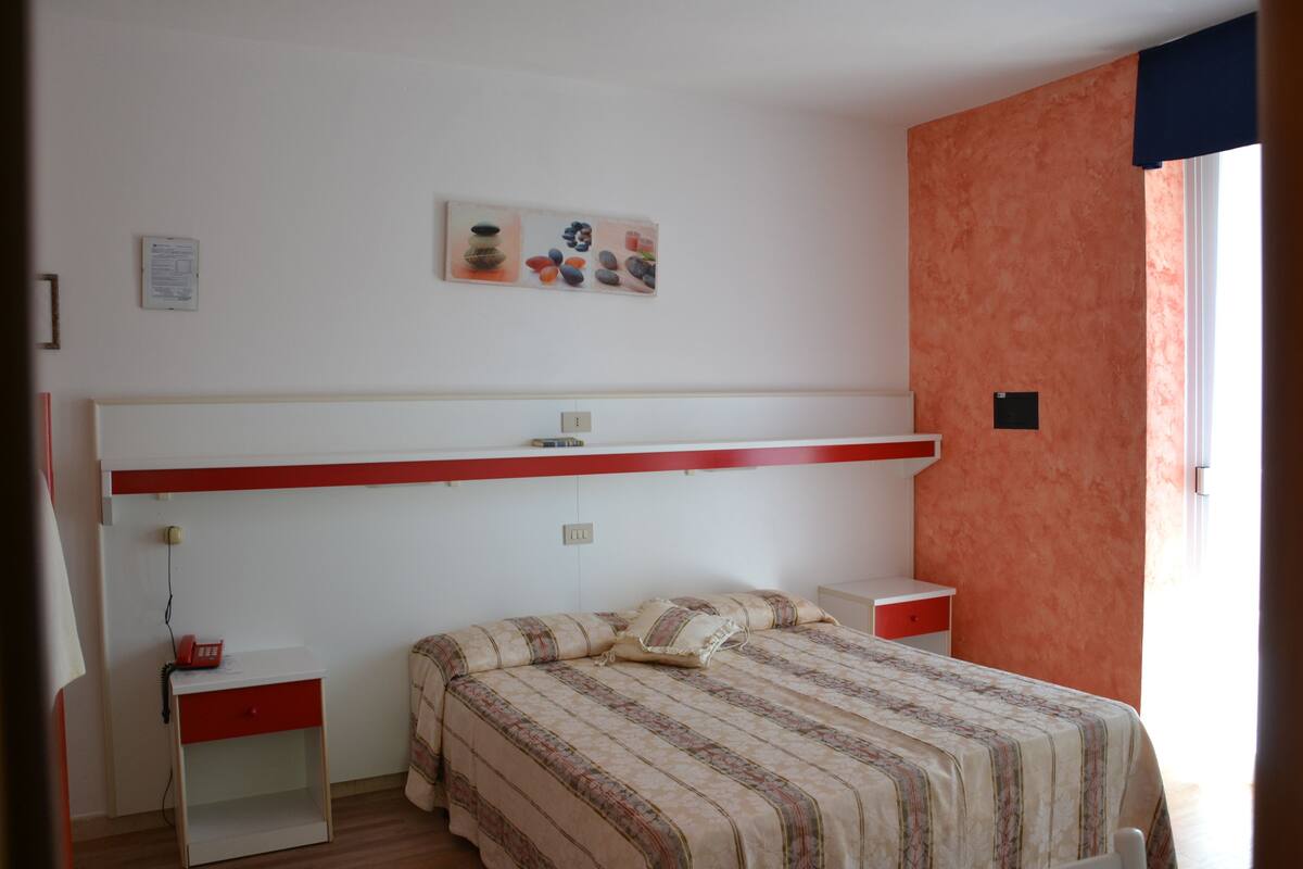 A bedroom with a bed, bedside tables, a nightstand, a wall-mounted shelf, and a window with curtains.