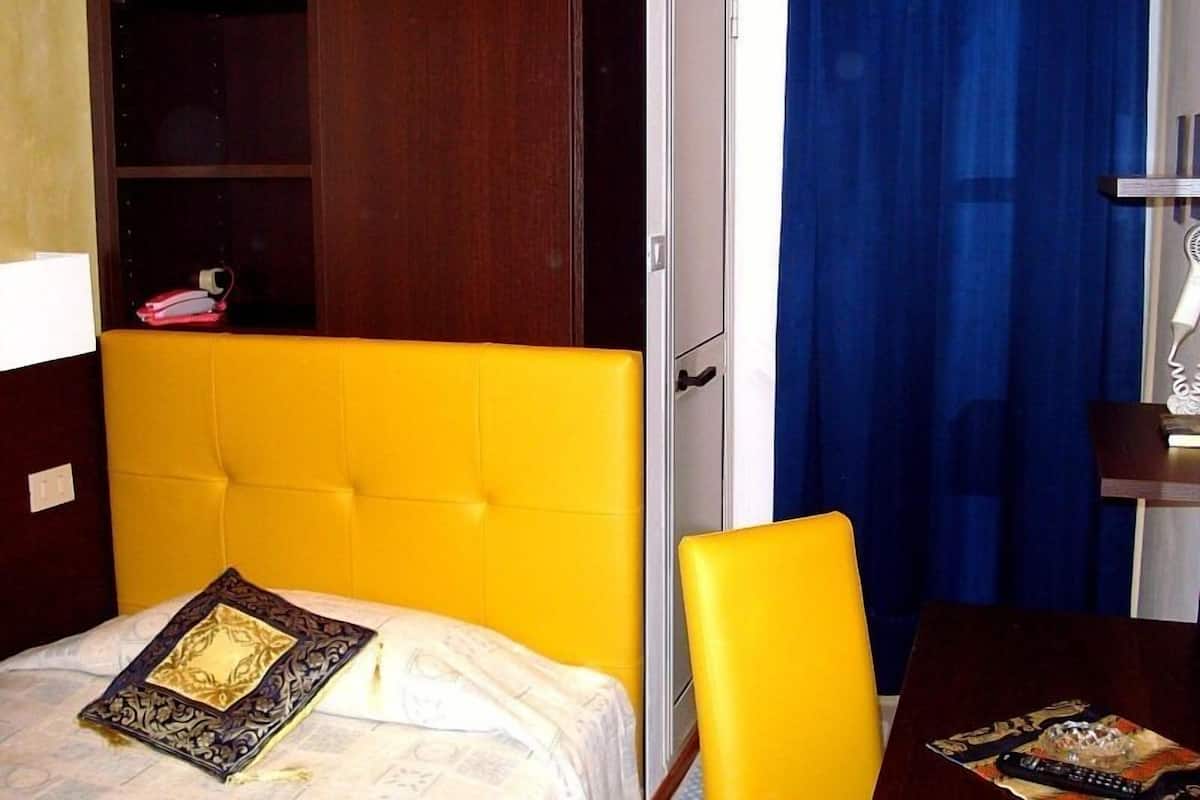 A room with a yellow headboard, a bed with a patterned pillow, a wooden wardrobe, a blue curtain, and a table with a remote control.