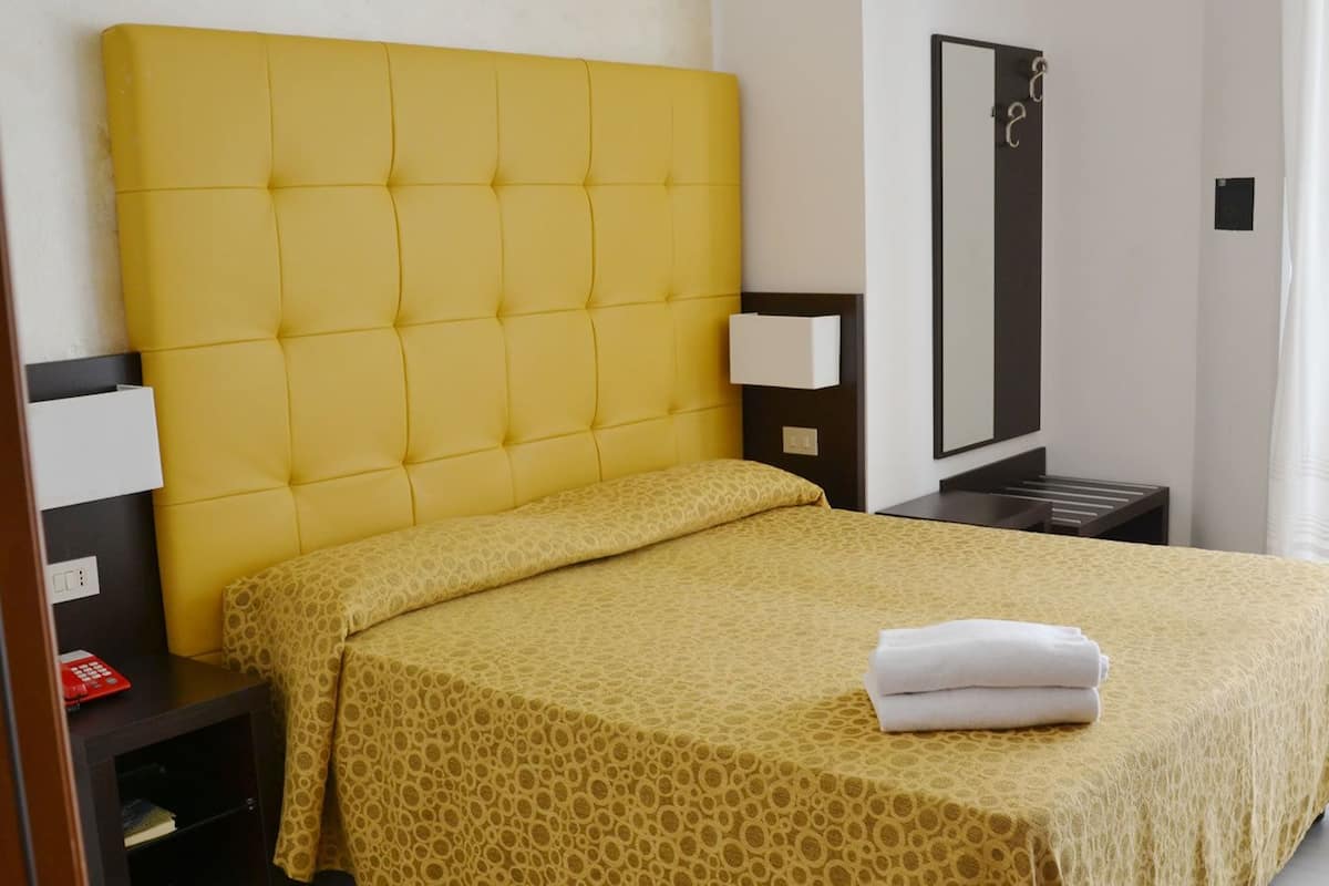 A hotel room with a yellow headboard, a bed with a patterned bedspread, a bedside table with a lamp, and a mirror on the wall.