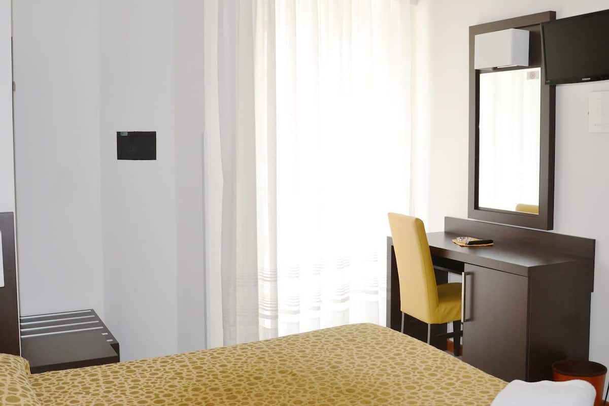 A hotel room with a bed, a desk, a chair, a mirror, and a window with curtains.