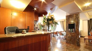 Uma área de recepção de hotel com balcão de madeira, mesa de atendimento, um pilar de mármore e um espaço de refeições com cadeiras e uma mesa.