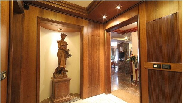 Corredor interno de madeira com uma estátua em pedestal, uma porta e um vislumbre de uma sala de jantar com mesa e cadeiras.