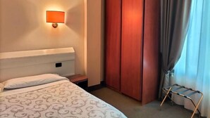 Quarto de hotel com cama, mesa de cabeceira, guarda-roupa de madeira, luminária na parede e janela com cortinas.
