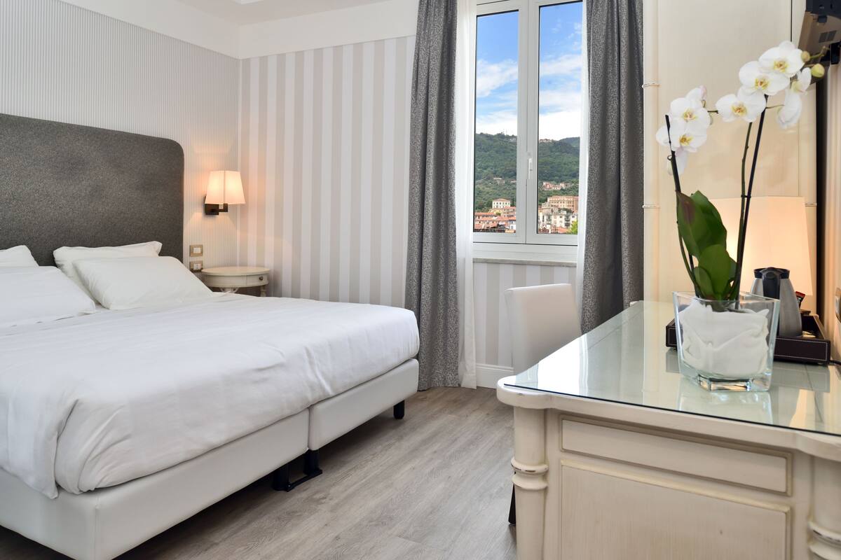 Hotelkamer met een bed, nachtkastje, lamp, glazen bijzettafeltje met bloevas en een raam met uitzicht op gebouwen en groen.