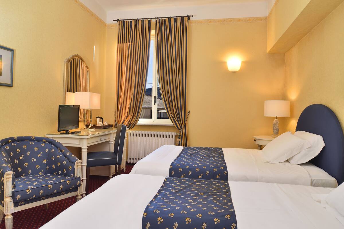 Een hotelkamer met twee bedden, een bureau met een computer, een stoel en een raam met gordijnen.