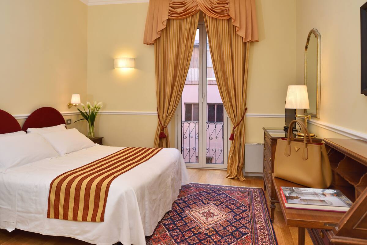 Hotelkamer met een bed, nachtkastje, lamp, dressoir en uitzicht door een raam met gordijnen.