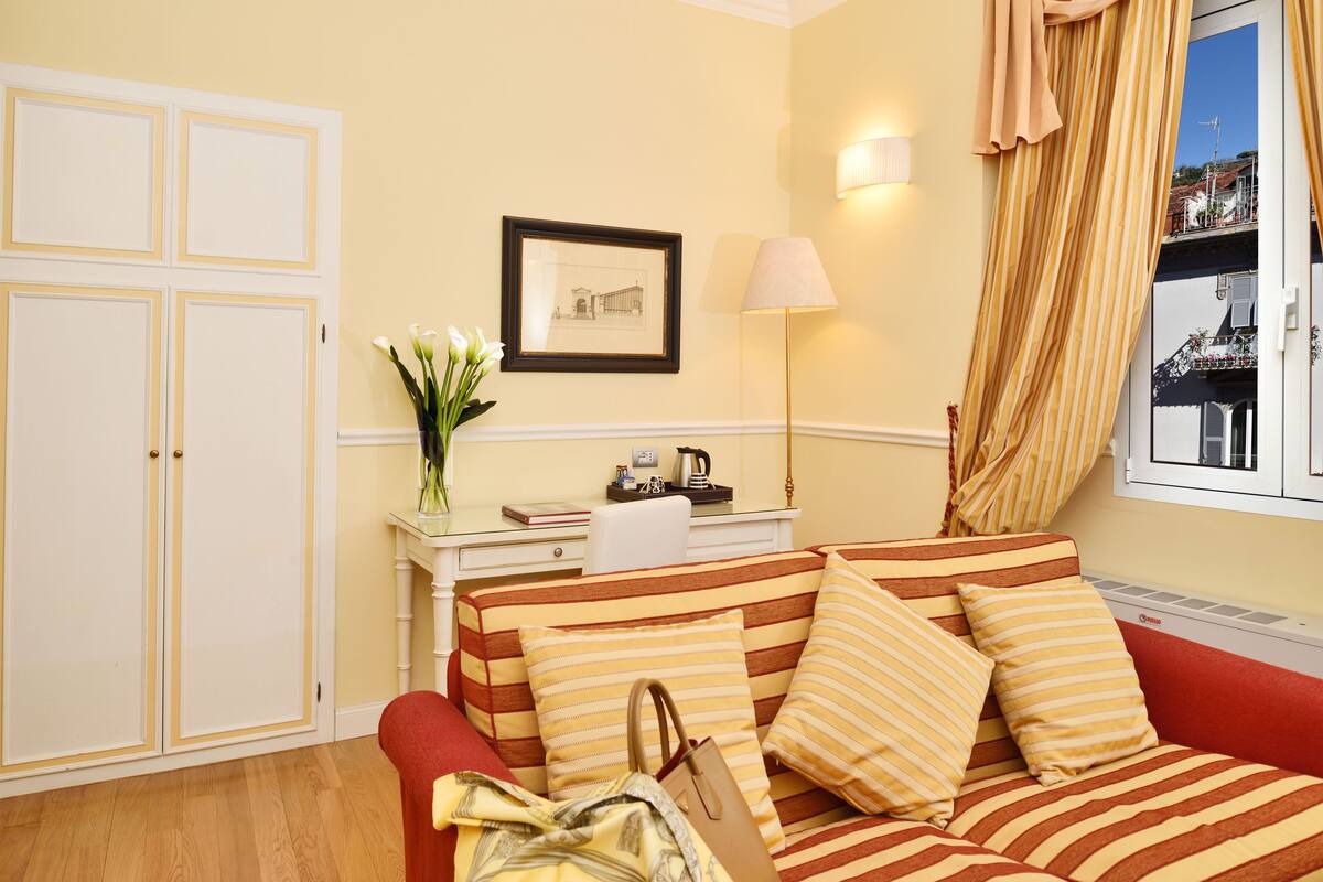 Een hotelkamer met een gestreepte bank, een bureau met een lamp, een ingelijst schilderij aan de muur en een raam met gordijnen.