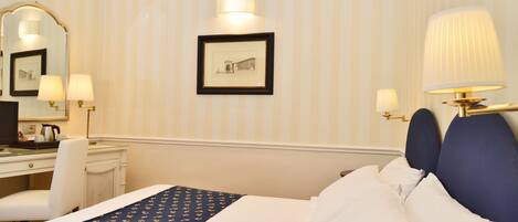 A hotel room with a bed, a desk, a chair, a lamp, and a framed picture on the wall.