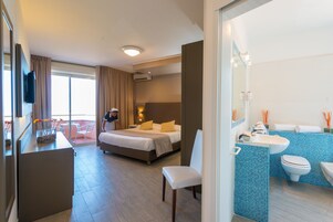 A hotel room with a bed, a desk, a chair, a balcony with a view, and a bathroom with a sink and toilet.