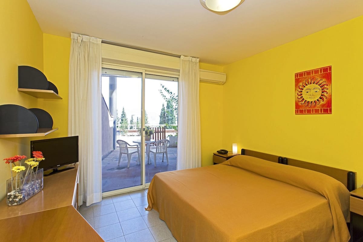 Habitación de hotel con cama, televisor, escritorio con un florero y puerta corrediza de vidrio a un balcón, además de una ilustración de sol en la pared.