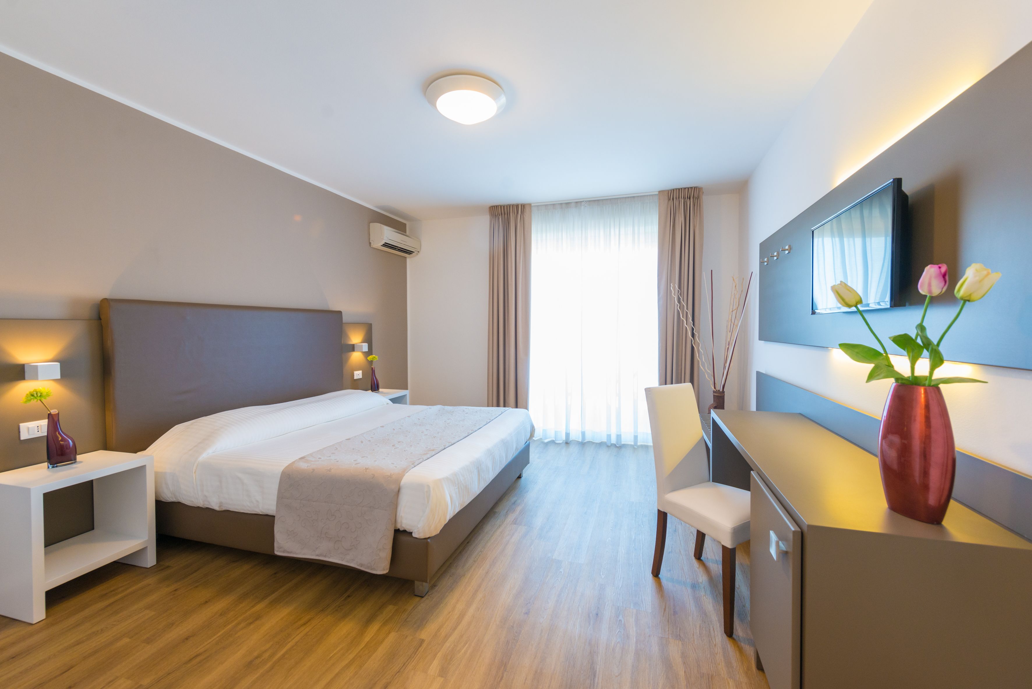 Moderni hotellihuone, jossa on sänky, yöpöydät, työpöytä, tuoli, televisio ja maljakko, jossa on kukkia.