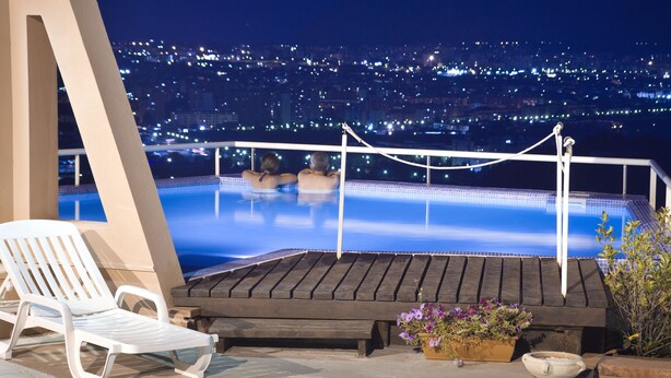 A rooftop pool area with a view of a cityscape at night, a lounge chair, and a wooden bench.