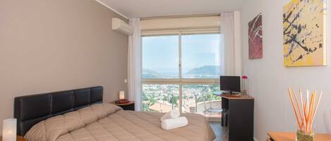A hotel room with a large bed, a desk with a computer, a window with a city view, and artwork on the wall.