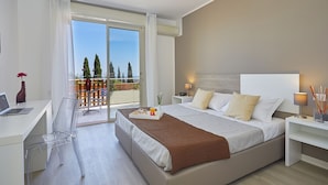A modern hotel room with a large bed, a desk with a laptop, a clear chair, and a balcony with a view of trees.