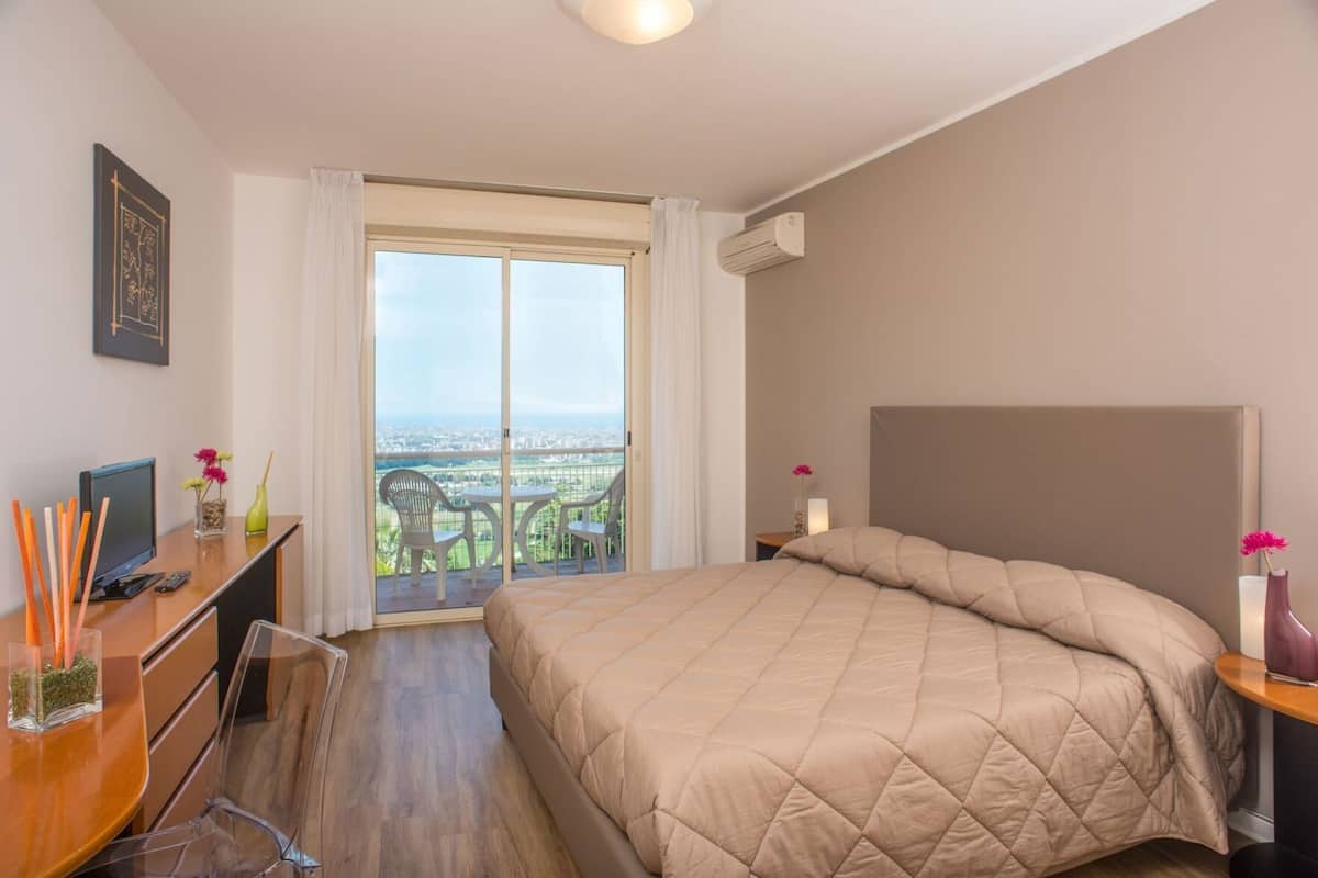 Un cuarto de hotel con una cama, un escritorio con una computadora, una silla, una mesita de noche y vistas a la ciudad a través de un ventanal grande.