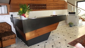 Uma mesa de recepção com acabamento em madeira, um vaso com planta e uma tela digital.