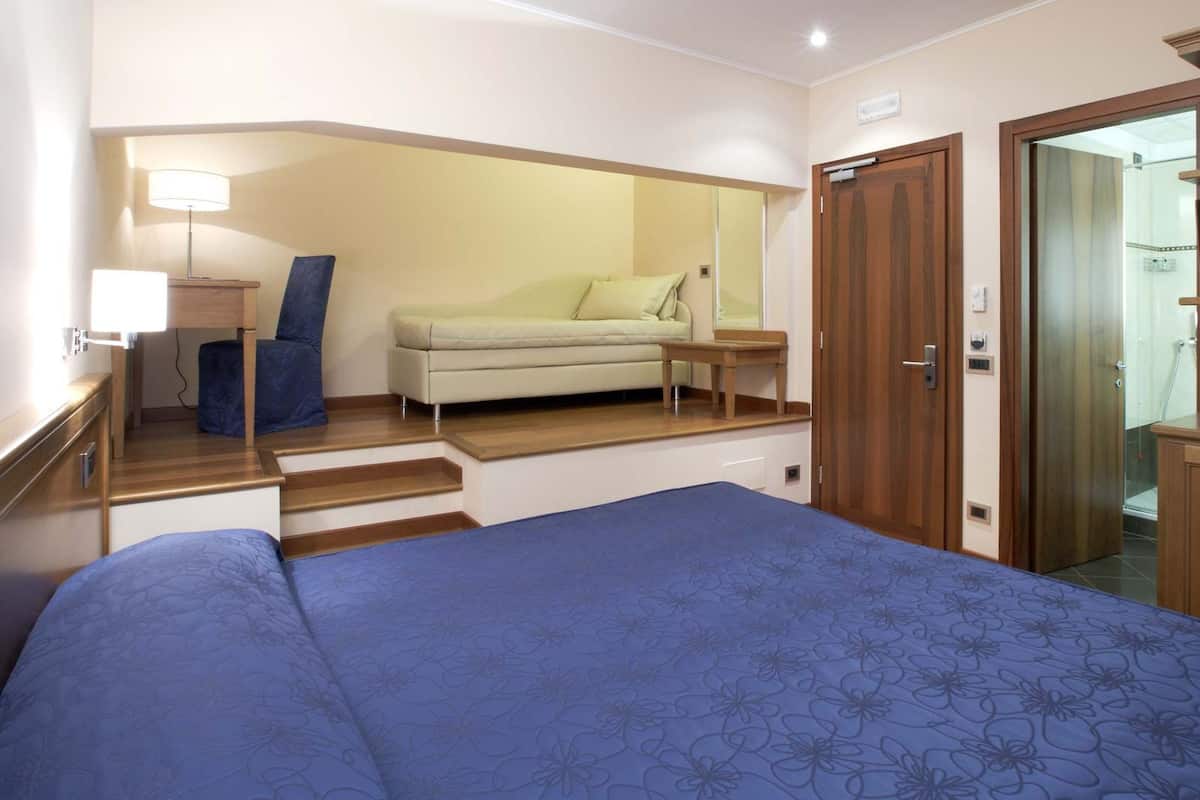 A bedroom with a bed, a sofa, a nightstand, a lamp, and a door leading to a bathroom.