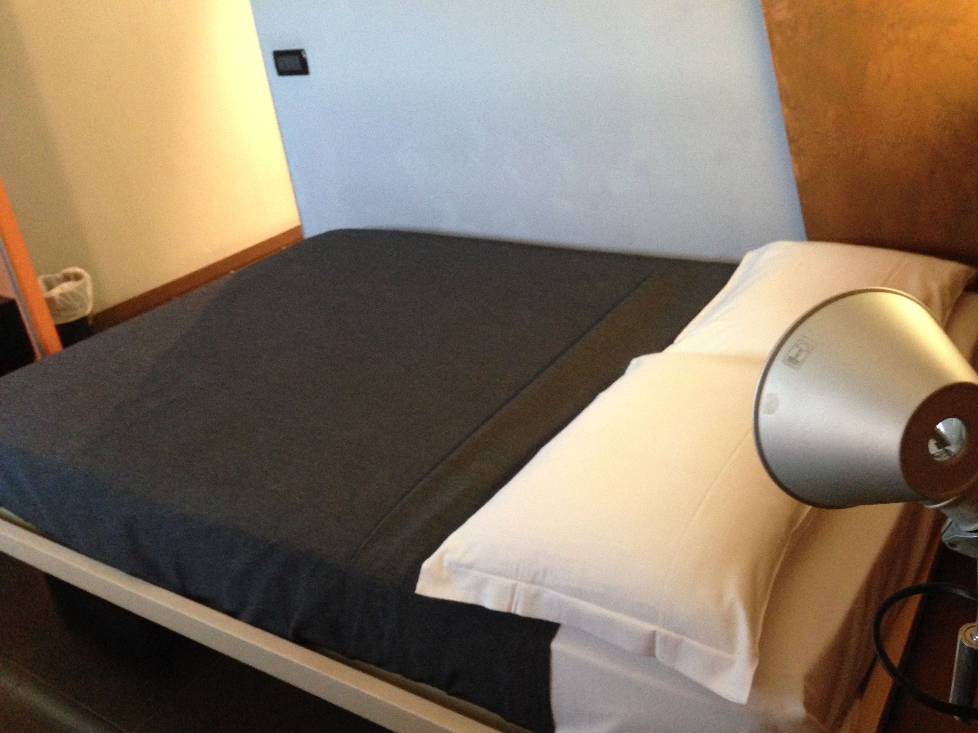 Ein Bett mit dunkler Bettdecke und weißen Kissen, ein Nachttisch mit Lampe sowie ein Holzkopfende.