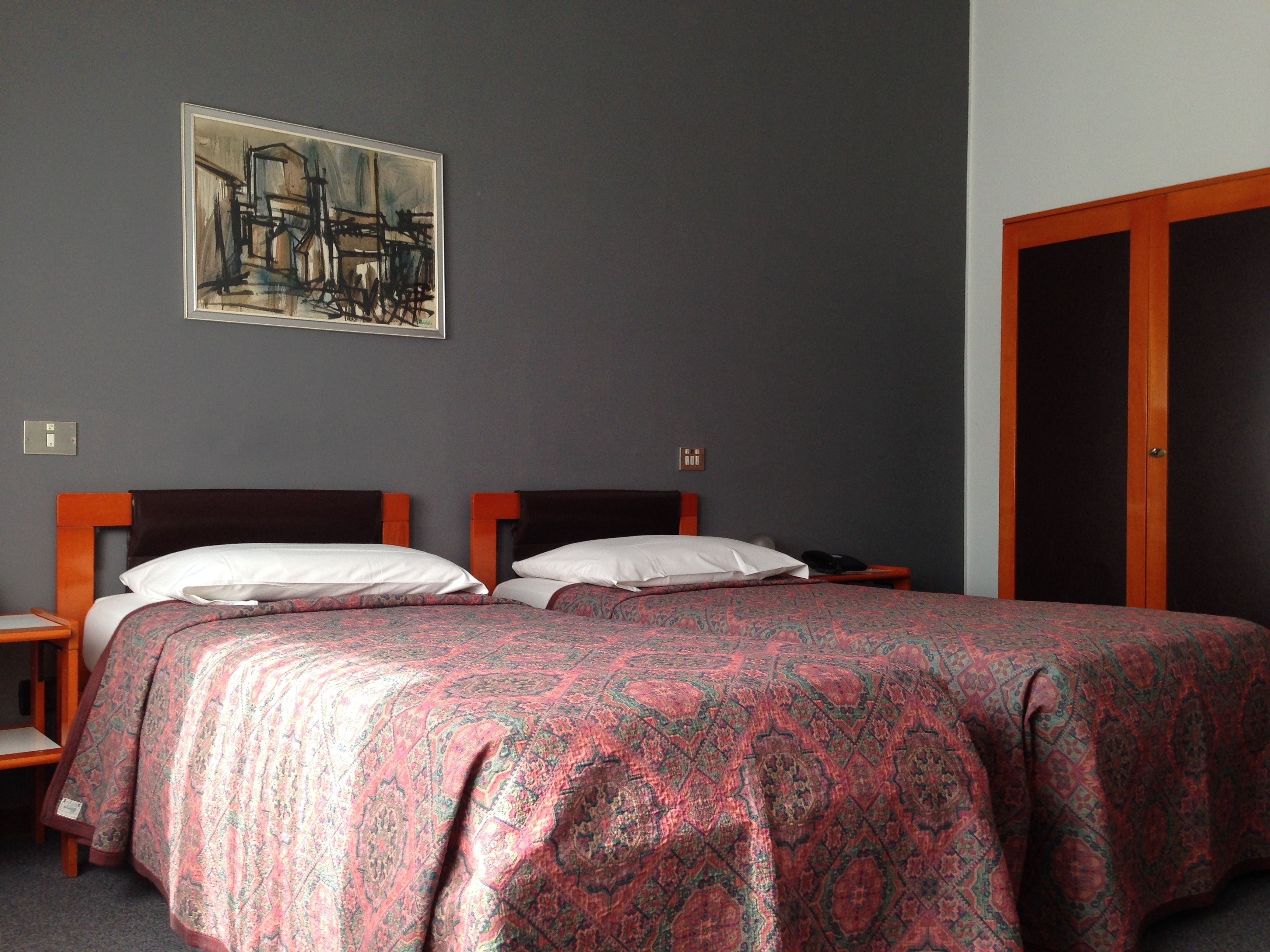 A double bed with a patterned bedspread, a nightstand, a wall-mounted painting, and a wooden wardrobe.