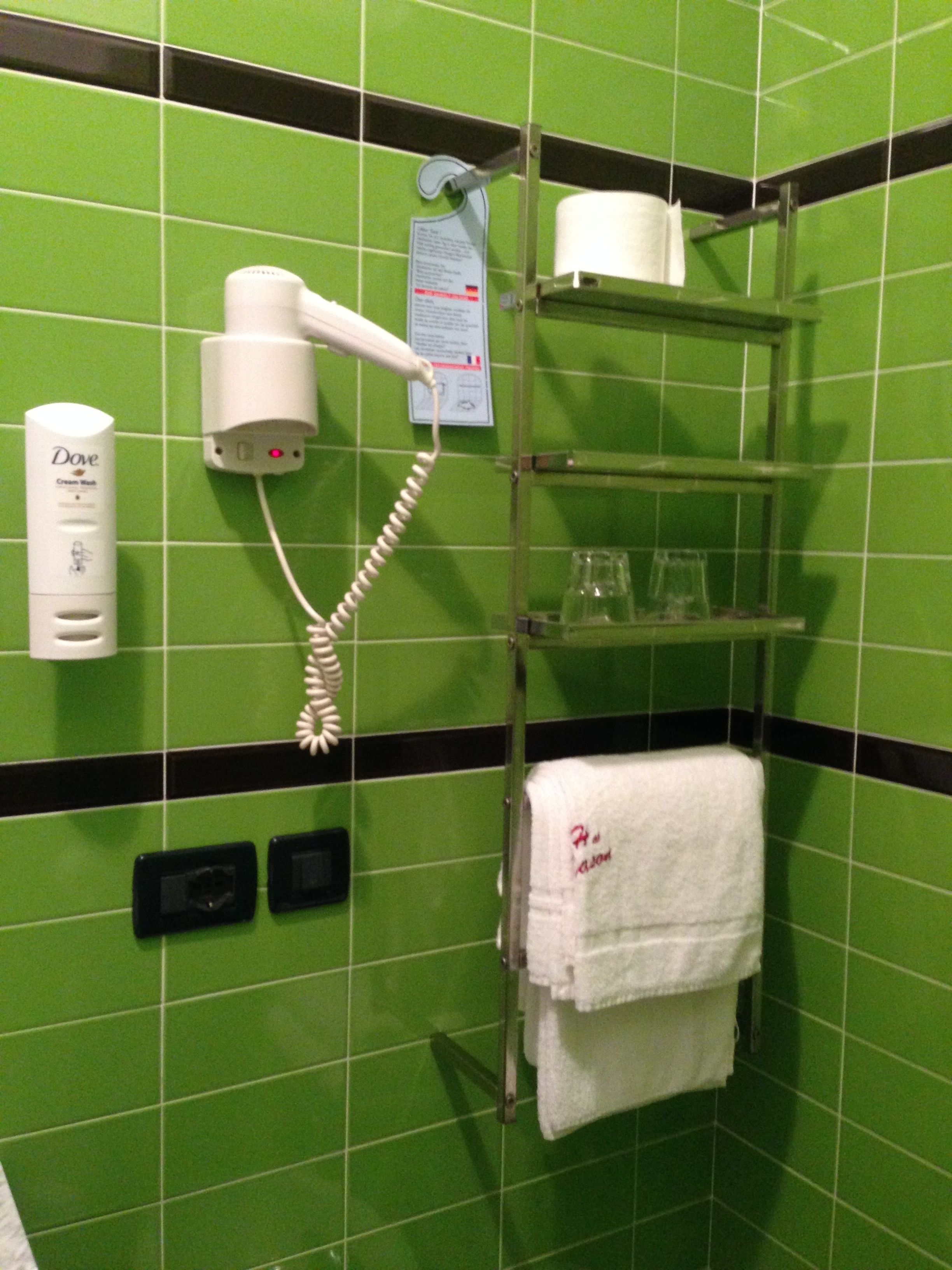 A bathroom with green tiled walls, a hairdryer, a Dove cream wash bottle, a towel rack with a towel, and a shelf with toiletries.