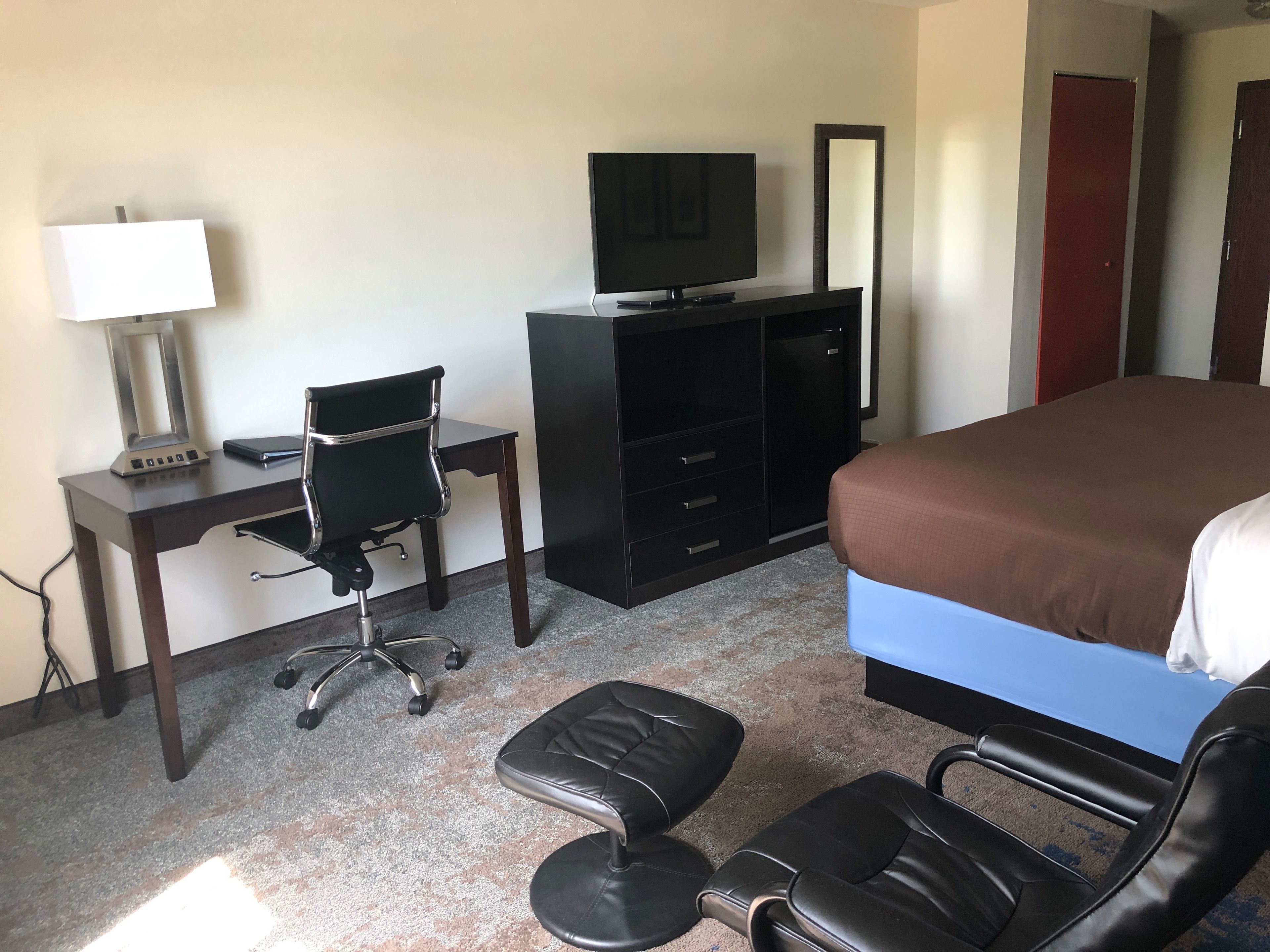 Habitación de hotel con cama, escritorio con silla, televisión, espejo y mesita de noche con lámpara.
