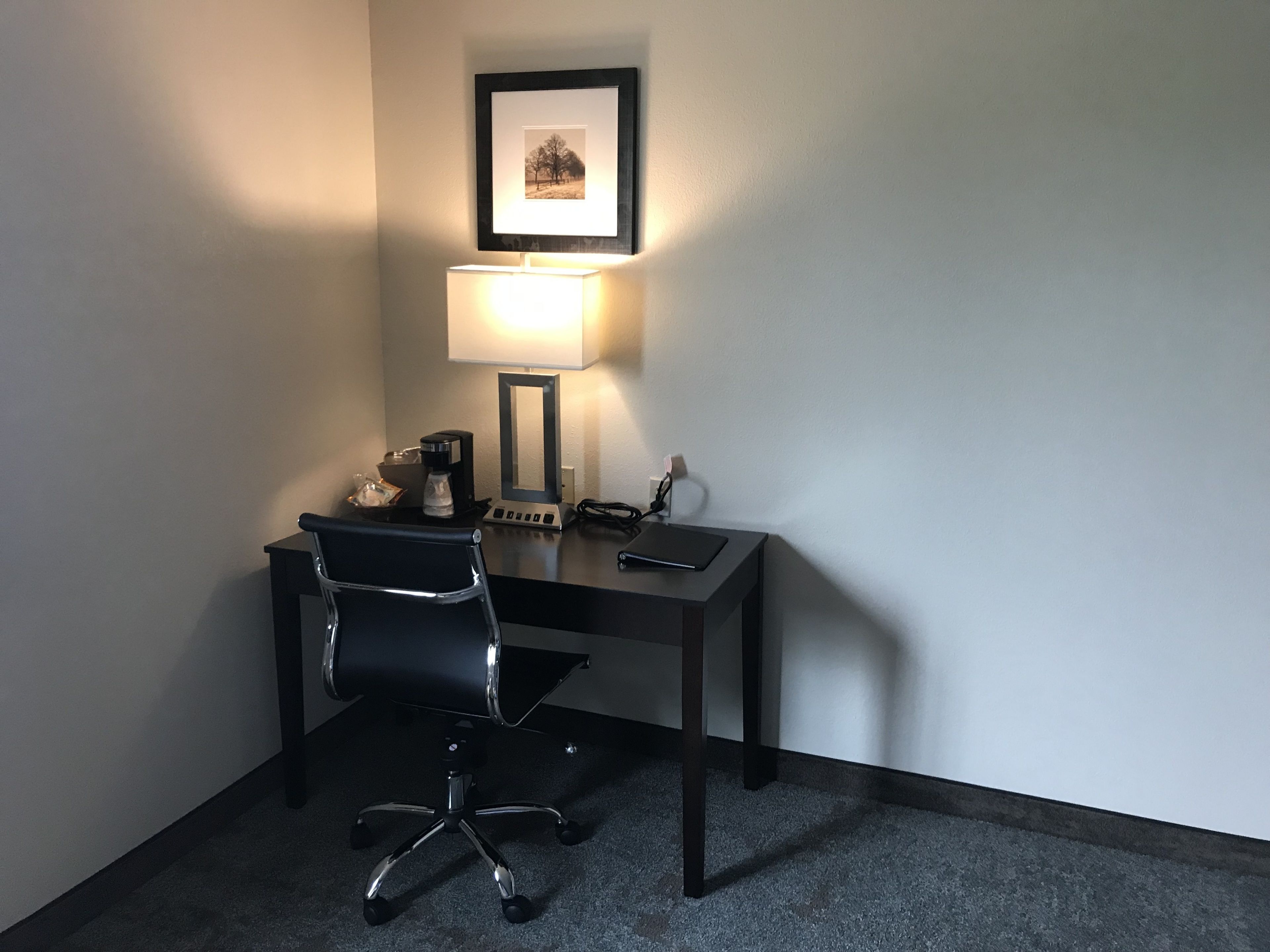 Een kleine kantoorruimte met een bureau, stoel, lamp en een ingelijst beeld aan de muur.