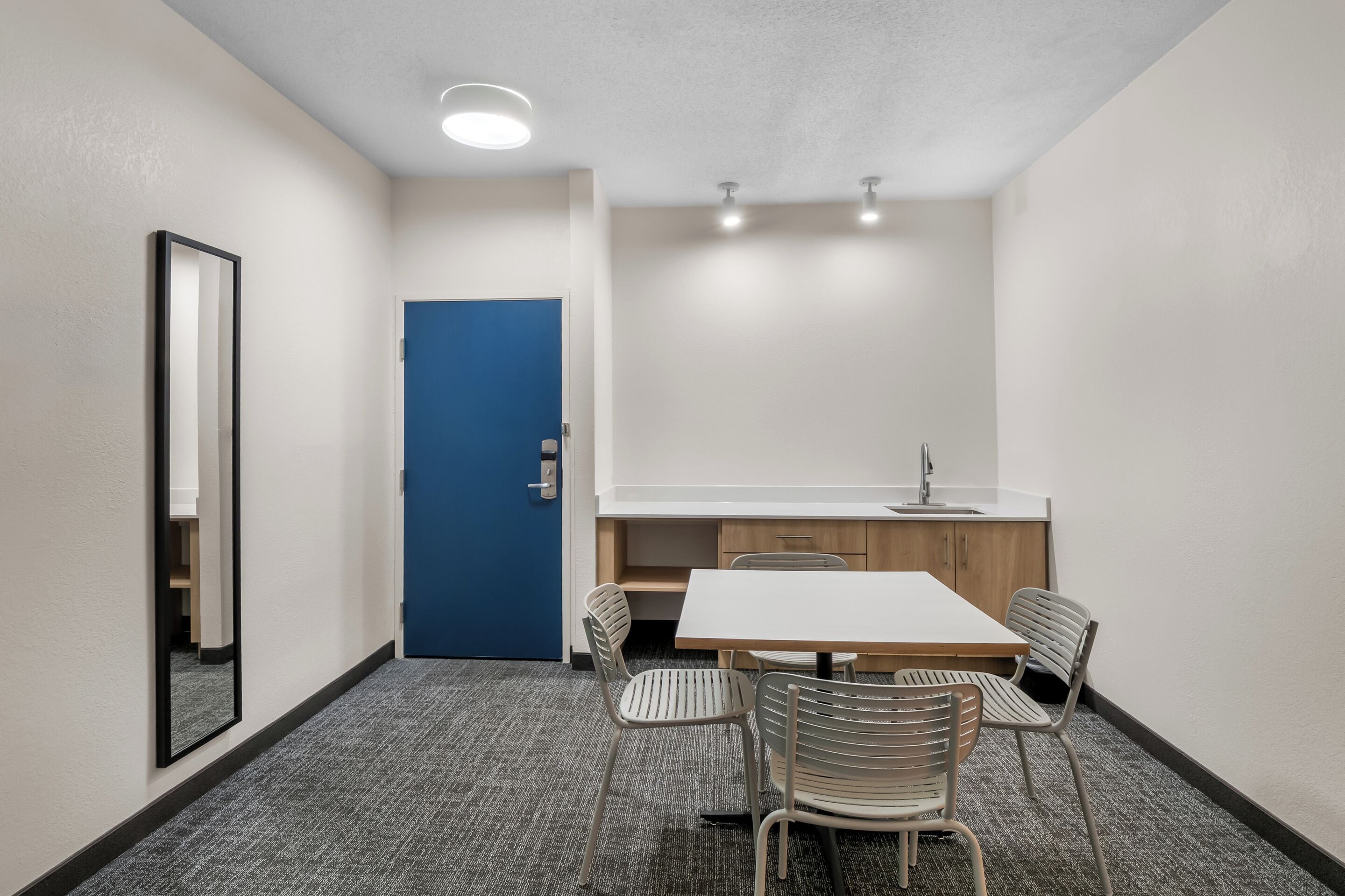 Una habitación pequeña y moderna con una puerta azul, una mesa blanca con cuatro sillas, una kitchenette y un espejo en la pared.