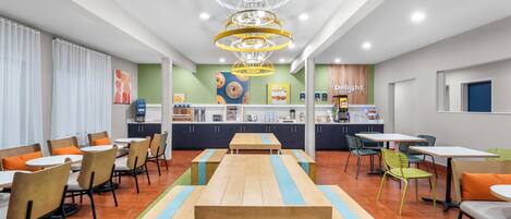A modern cafe with wooden tables and chairs, a central counter with a coffee machine, and a green wall with artwork.