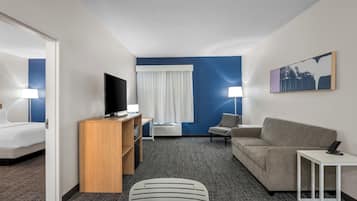 A hotel room with a flat-screen TV, a sofa, a chair, a small table, a desk, a bookshelf, and a window with curtains.