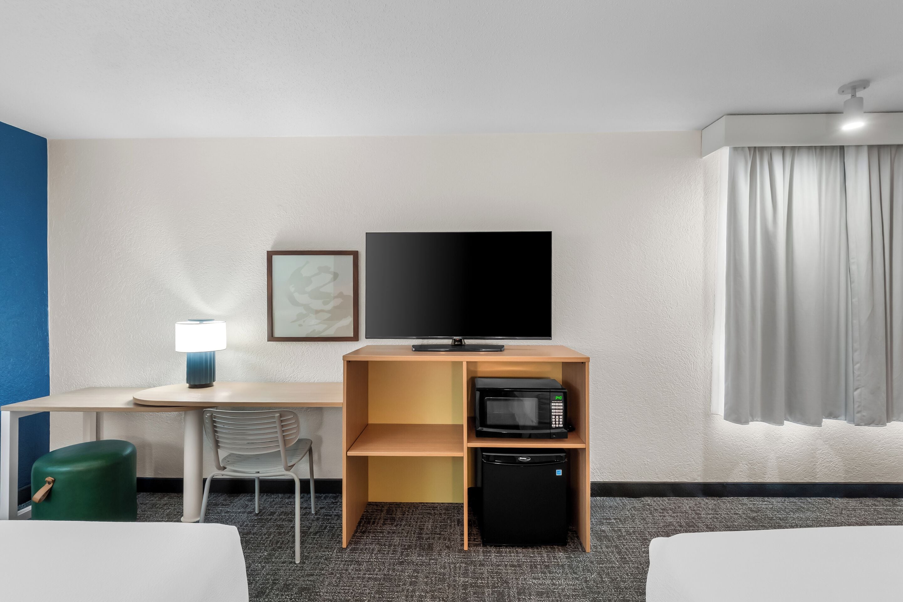 A hotel room with a bed, a desk with a lamp, a television, a microwave, and a refrigerator.