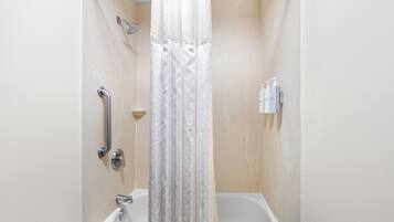 A bathroom with a shower, a handrail, a bathtub, and a wall-mounted soap dispenser.