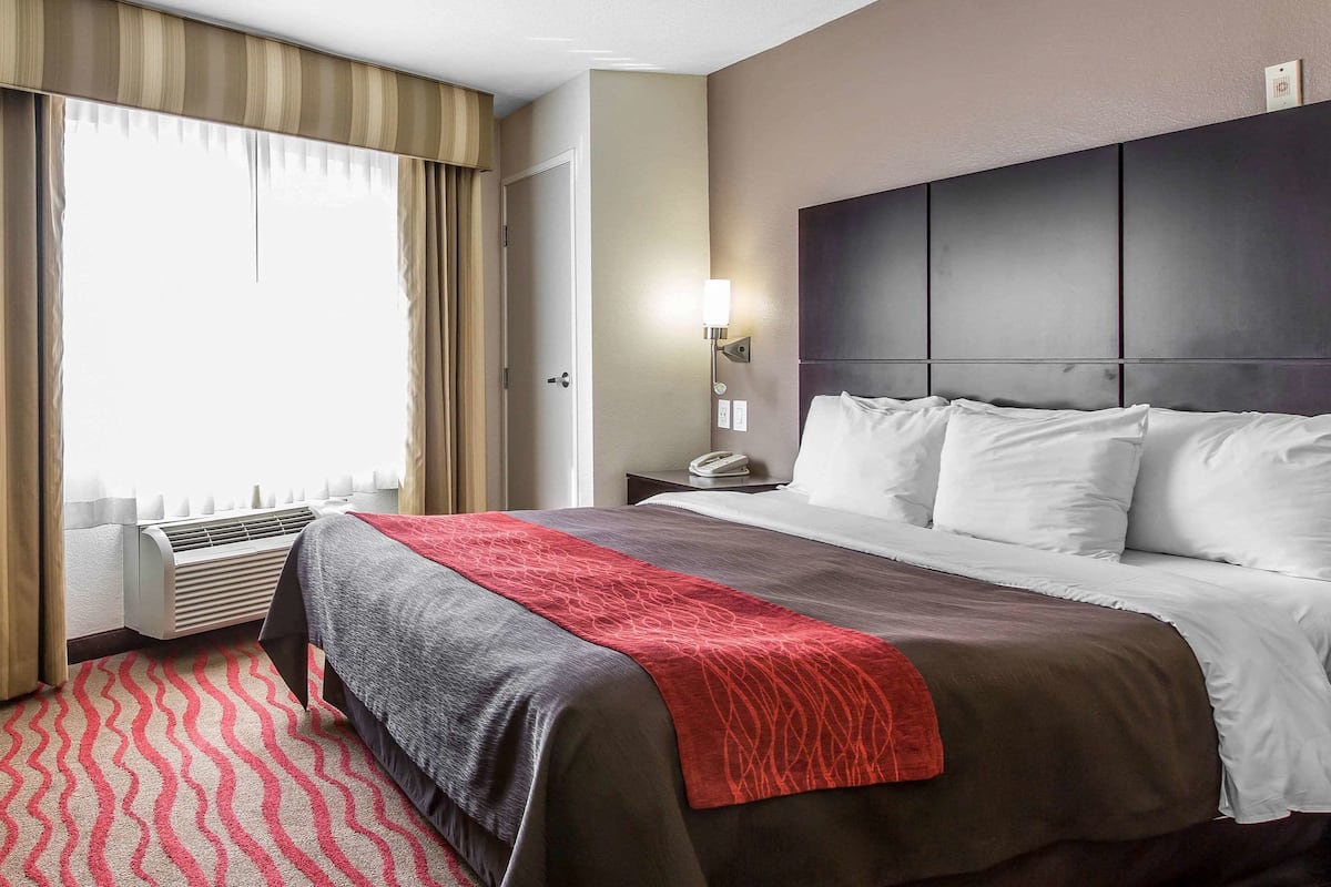 A hotel room with a large bed, a bedside table with a lamp, a window with curtains, and a wall-mounted air conditioning unit.