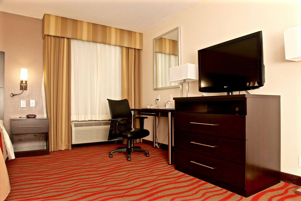 A hotel room with a large flat-screen TV, a desk with a chair, a dresser, and a red carpeted floor.