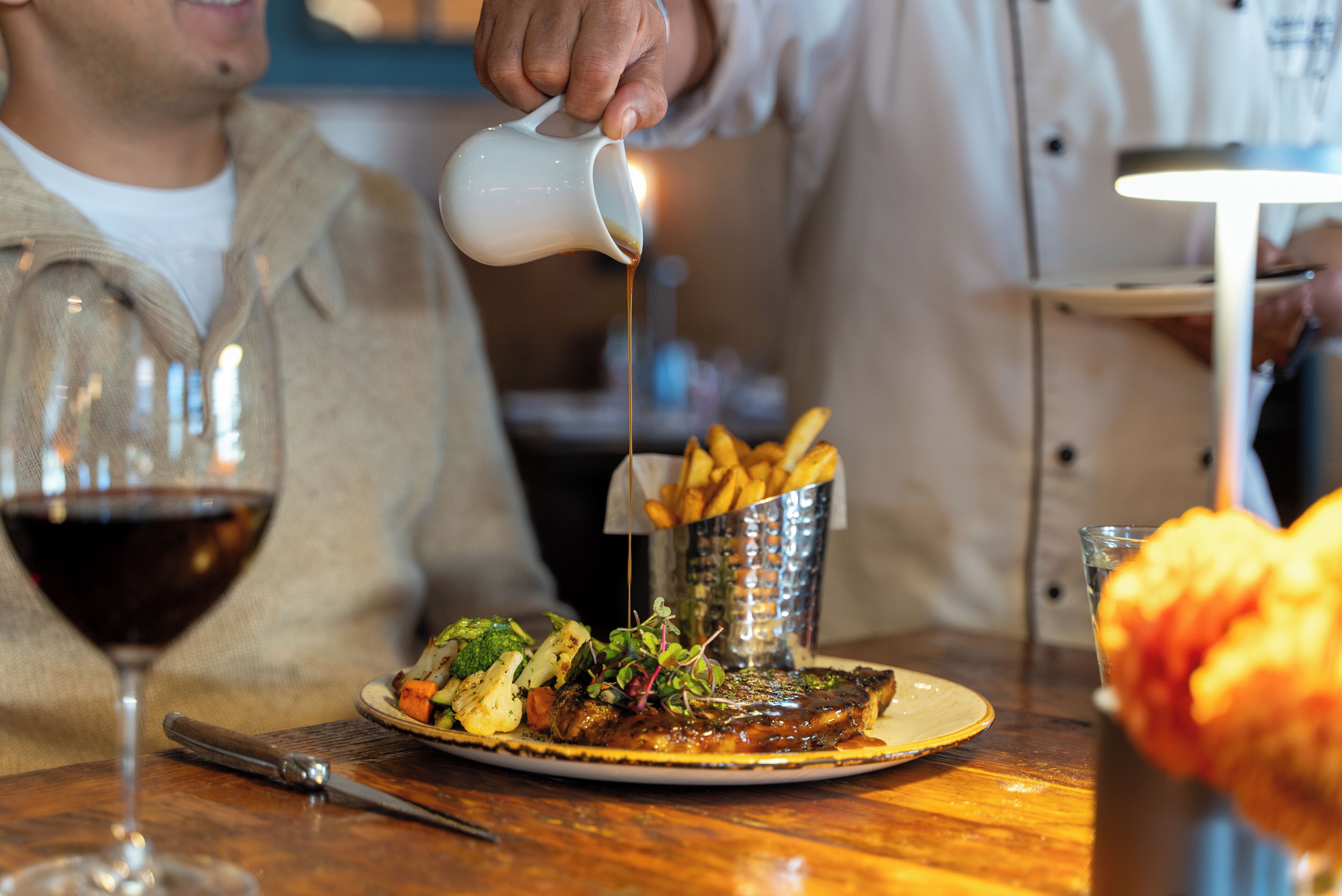 Ein Esstisch mit einem Teller Essen, einem Glas Rotwein und einer Metallbox mit Pommes.