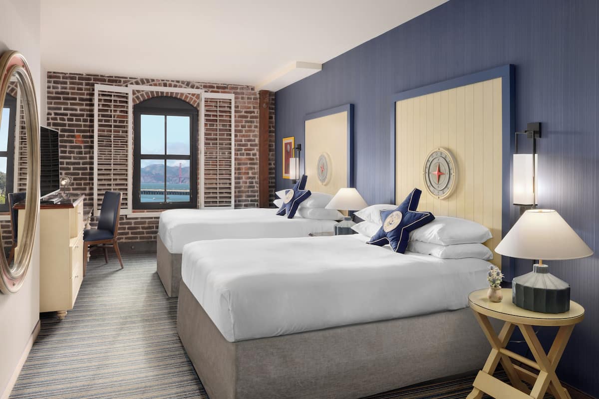 Een hotelkamer met twee bedden, een bakstenen muur, een groot raam en uitzicht op een brug.