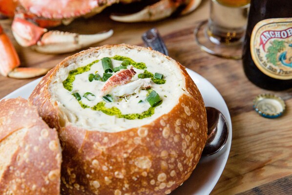 Un bol de soupe servi dans un bol en pain, accompagné d’une cuillère, une bouteille de bière Anchor et un crabe posés sur la table.