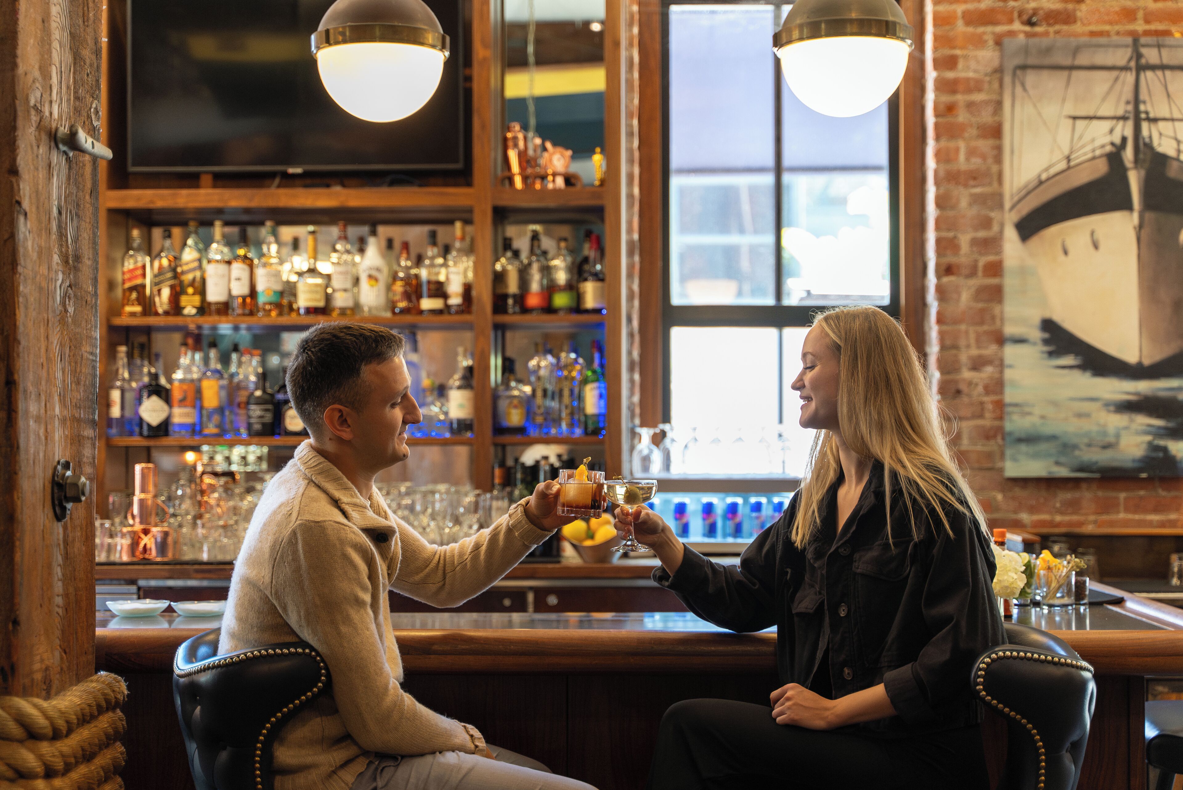 A bar with wooden shelves stocked with bottles, a large window, and a ship painting on the wall.