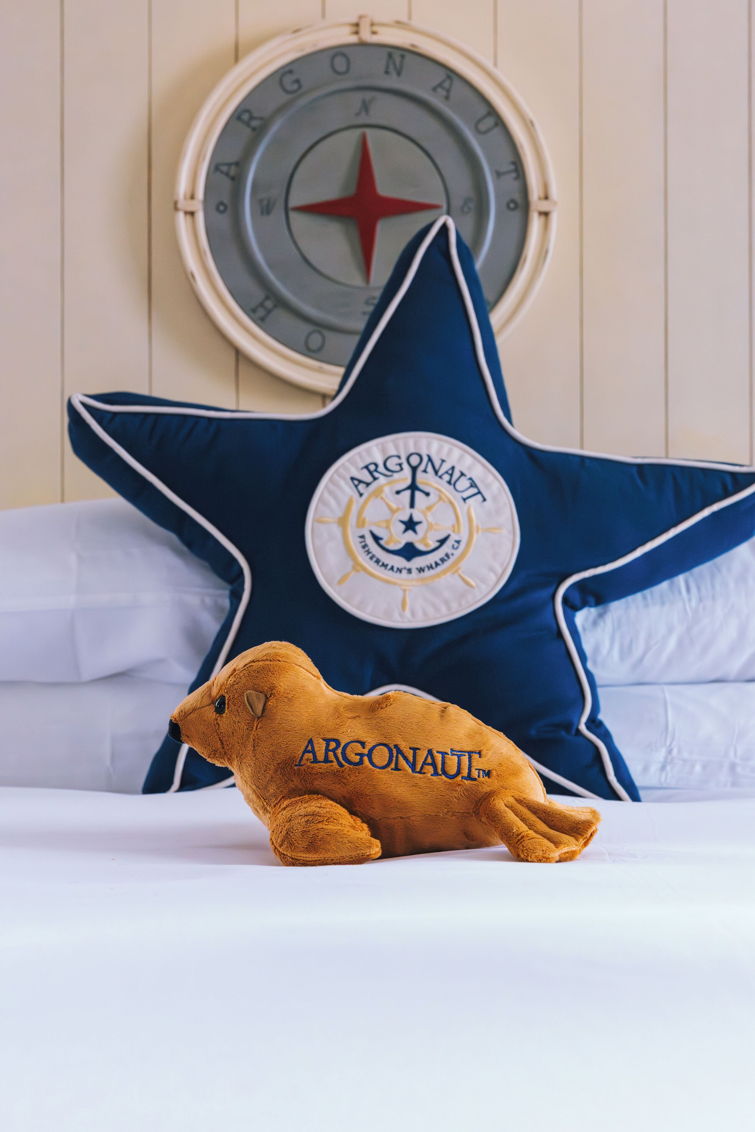 A bed with a large blue star-shaped pillow featuring the Argonaut logo, a compass wall clock, and a plush toy with the Argonaut logo.