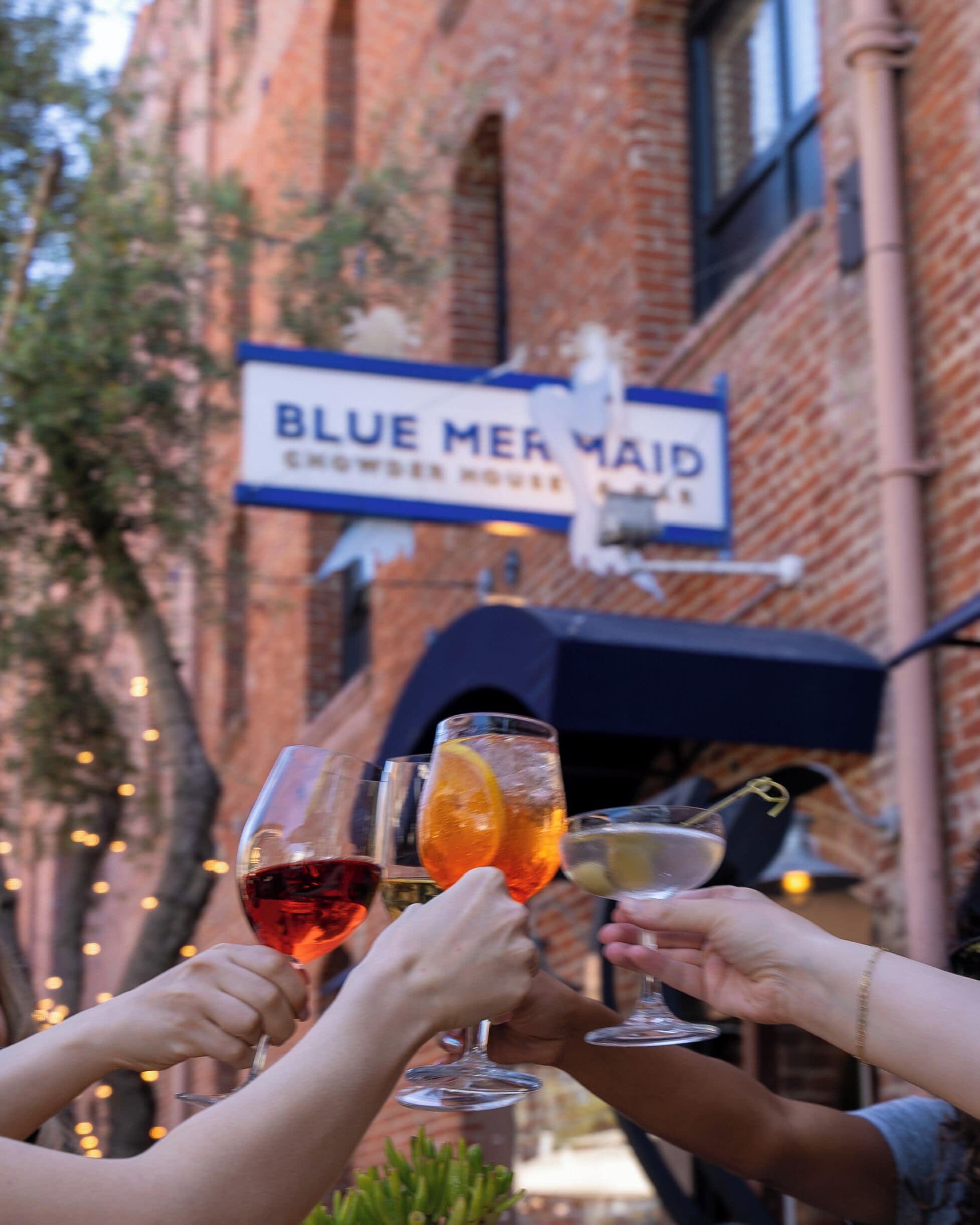 A group of people toasting with drinks in front of a brick building with a sign that reads "BLUE MERMAID".