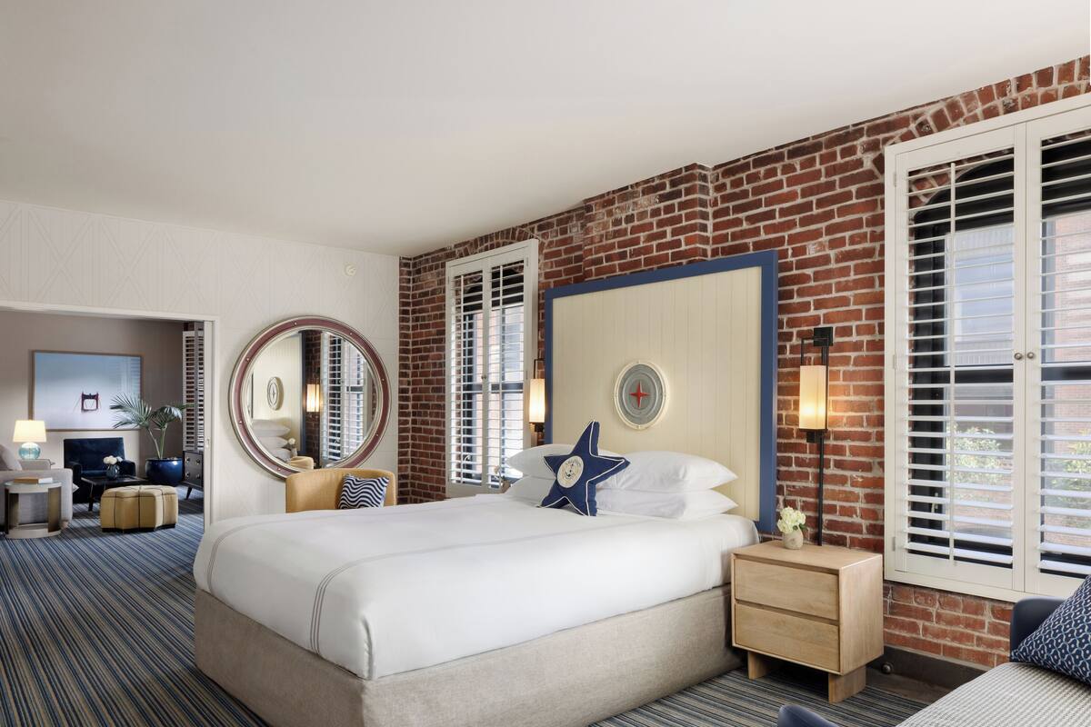 Een hotelkamer met een groot bed, een ronde spiegel, een bureau met een lamp en uitzicht buiten door een raam met jaloezieën.